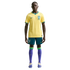A full-length studio portrait of a man modeling the Brazil national football team uniform against a solid black background. He is wearing the team's iconic yellow jersey with green accents, blue shorts, purple socks with green bands at the top, and light