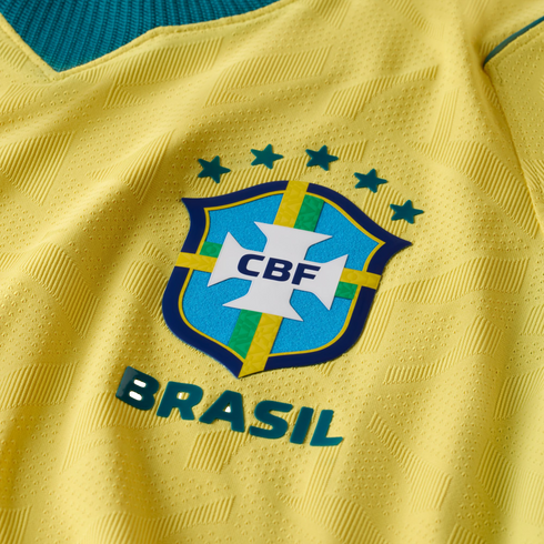 A close-up of a yellow Brazilian national football team jersey, featuring the blue CBF crest, five teal stars above it, and the word "BRASIL" in raised letters below. The yellow fabric has an intricate, textured geometric pattern.