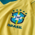 A close-up of a yellow Brazilian national football team jersey, featuring the blue CBF crest, five teal stars above it, and the word "BRASIL" in raised letters below. The yellow fabric has an intricate, textured geometric pattern.