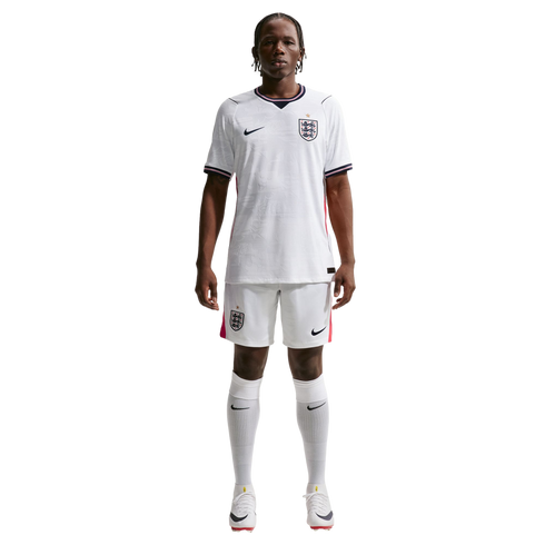 A full-body photograph of a male athlete standing against a solid black background, wearing the white Nike England national team home soccer kit. The kit includes a white jersey and shorts featuring the team's Three Lions crest and Nike logo, paired with