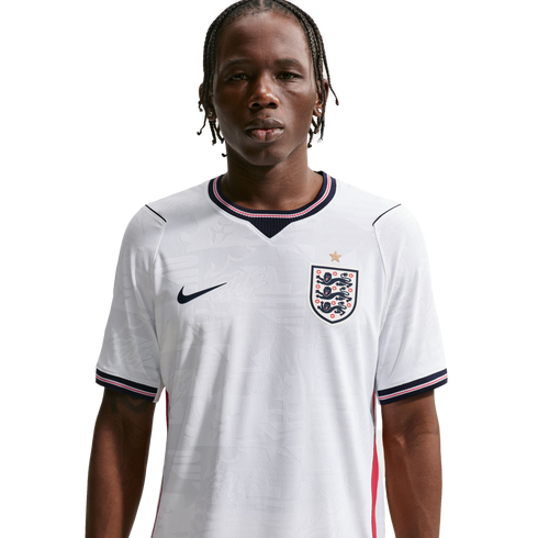 A waist-up portrait of a young man with braided hair wearing a white Nike England national football team home jersey against a solid black background.