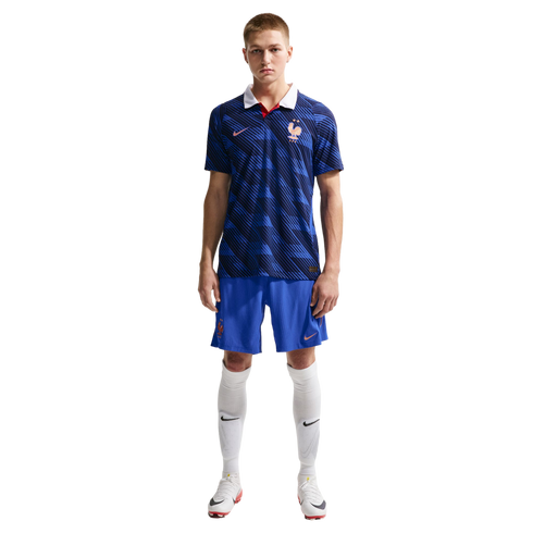 A man in a blue patterned France national football kit with a white collar, blue shorts, and white socks, standing against a black background.