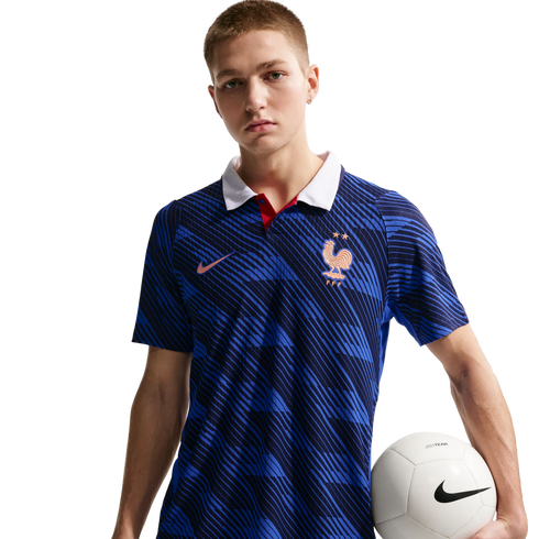 A man wearing a blue Nike France national team soccer jersey with a white collar, holding a white soccer ball against a black background.