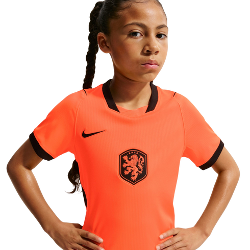 A young girl with a long braid and a serious expression poses with hands on her hips, wearing an orange Nike Netherlands national team soccer jersey against a solid black background.