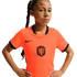 A young girl with a long braid and a serious expression poses with hands on her hips, wearing an orange Nike Netherlands national team soccer jersey against a solid black background.