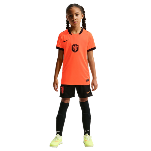 A young girl stands facing forward in a full Netherlands national soccer kit, including a bright orange Nike jersey, black shorts, tall black socks, and neon yellow cleats, against a solid black background.