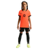 A young girl stands facing forward in a full Netherlands national soccer kit, including a bright orange Nike jersey, black shorts, tall black socks, and neon yellow cleats, against a solid black background.