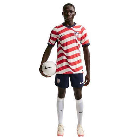 A male soccer player in a wavy red and white USA jersey and navy shorts, holding a soccer ball under his arm against a black background.