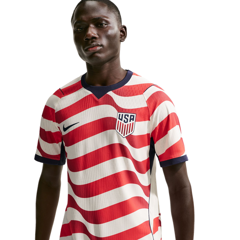 A man wearing a USA soccer jersey with red and white wavy stripes, featuring a Nike swoosh and USA crest, against a solid black background.