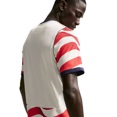 Rear view of a man looking down, wearing a white soccer jersey with red and white striped sleeves and navy blue trim against a black background.