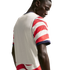Rear view of a man looking down, wearing a white soccer jersey with red and white striped sleeves and navy blue trim against a black background.
