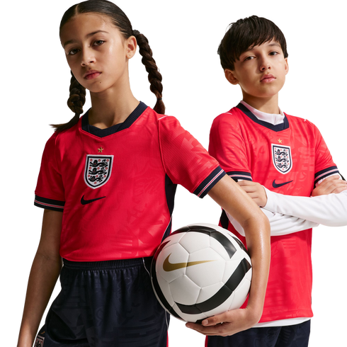 A girl and boy in red England football kits pose against a black background. The girl holds a Nike soccer ball while the boy stands with his arms crossed.