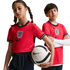 A girl and boy in red England football kits pose against a black background. The girl holds a Nike soccer ball while the boy stands with his arms crossed.