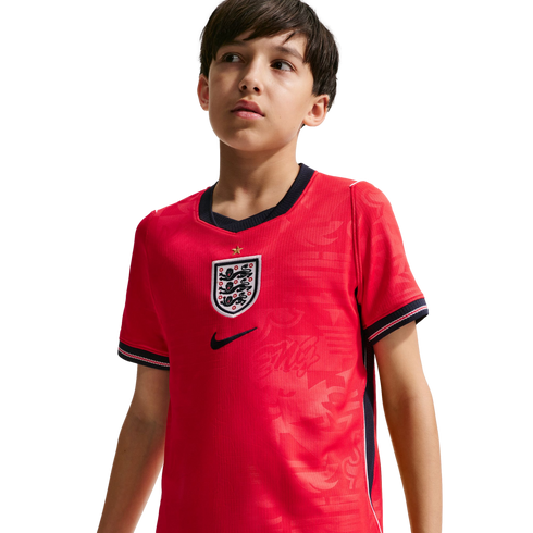 A young boy wearing a red England national football team jersey with the Three Lions crest, looking off-camera against a black background.