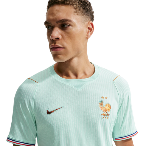 A man wears a mint green France national soccer team jersey with a golden rooster crest and tricolor sleeve cuffs, looking away against a black background.
