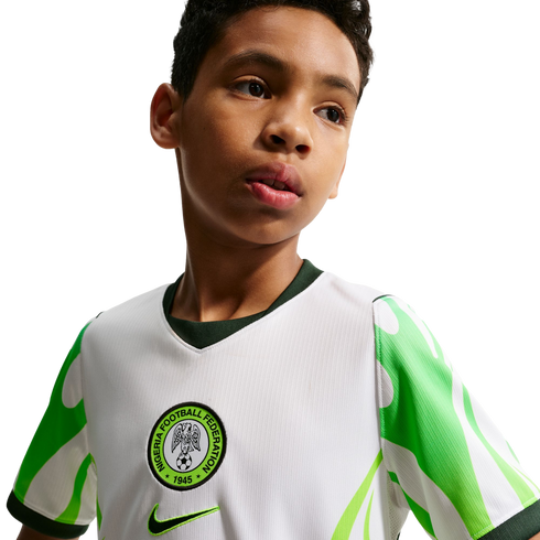 A young boy looking off-camera, wearing a white Nigeria national football team jersey with green patterned sleeves and the federation's crest.