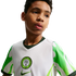 A young boy looking off-camera, wearing a white Nigeria national football team jersey with green patterned sleeves and the federation's crest.