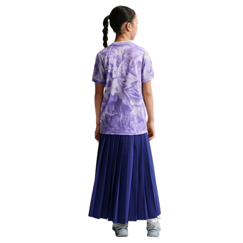 Back view of a girl with a braided ponytail wearing a purple floral t-shirt and a long navy pleated skirt against a black background.