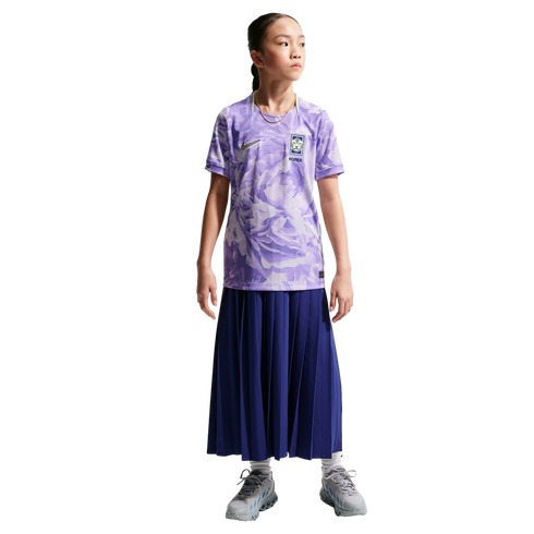 A young girl in a purple floral Nike South Korea soccer jersey, long navy pleated skirt, and gray sneakers, standing against a black background.