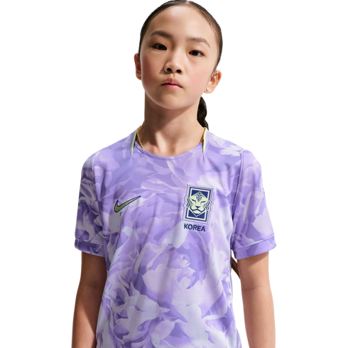 Portrait of a young girl wearing a light purple patterned Nike South Korea national team soccer jersey against a solid black background.