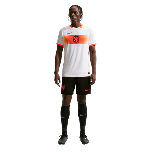 A full-length portrait of a male athlete wearing a Netherlands national soccer team away kit, standing against a solid black background. He is wearing a white jersey with an orange horizontal stripe across the chest, black shorts, and black knee-high sock