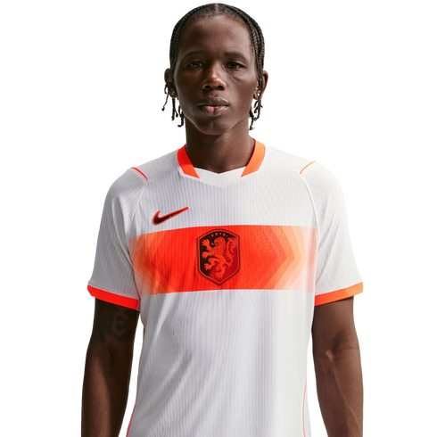 A young man with braided hair wears a white Netherlands national football team away jersey with a horizontal orange patterned band, the KNVB lion crest, and a black Nike swoosh, set against a solid black background.