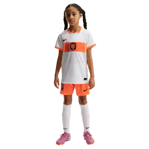 A young girl with braided pigtails stands against a black background, wearing a white and orange Nike Netherlands national team soccer kit, white socks, and pink cleats.
