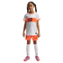 A young girl with braided pigtails stands against a black background, wearing a white and orange Nike Netherlands national team soccer kit, white socks, and pink cleats.