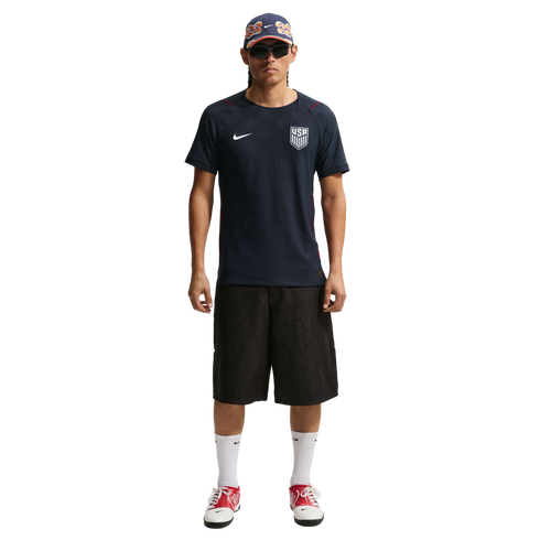 A man in a navy USA soccer jersey, black shorts, and red sneakers stands against a black background wearing a patterned cap and sunglasses.