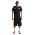 A man in a navy USA soccer jersey, black shorts, and red sneakers stands against a black background wearing a patterned cap and sunglasses.