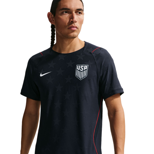 A man wearing a navy blue Nike USA soccer training jersey with star patterns and red accents, looking away from the camera against a black background.