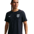 A man wearing a navy blue Nike USA soccer training jersey with star patterns and red accents, looking away from the camera against a black background.
