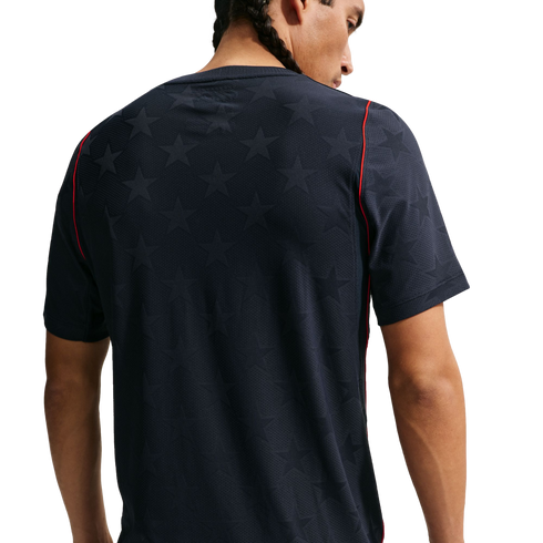 Back view of a man with braided hair wearing a dark navy t-shirt featuring a subtle star pattern and red piping on the shoulders, set against a plain black background.