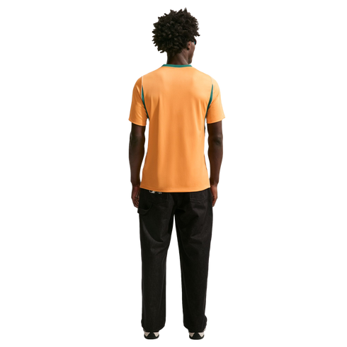 Back view of a person with dark curly hair wearing an orange shirt with green trim and black pants against a black background.