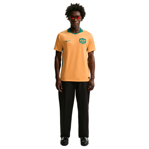 A man with curly hair and red sunglasses wears an orange and green Australia soccer jersey with black pants, standing against a solid black background.