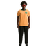 A man with curly hair and red sunglasses wears an orange and green Australia soccer jersey with black pants, standing against a solid black background.