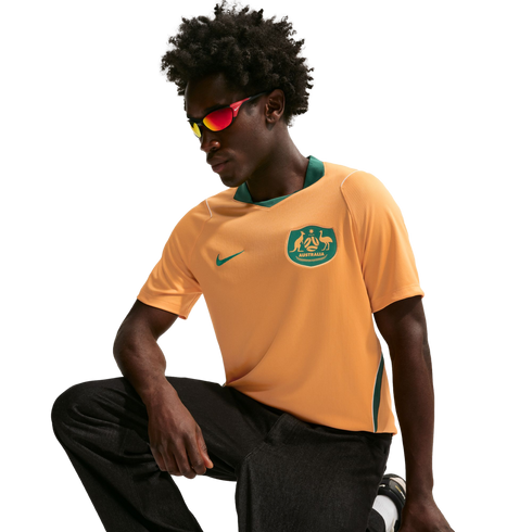 A man wearing an orange and green Australia national soccer jersey and red-tinted sunglasses crouches against a black background.
