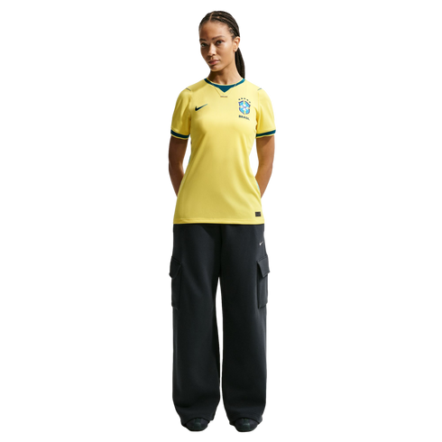 A woman with braided hair wearing a yellow Brazil national soccer team jersey and dark cargo pants, standing against a black background.