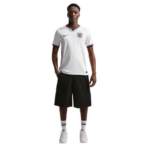 A man stands against a black background wearing a white England soccer jersey, black shorts, and white sneakers.