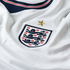 Close-up of a white England national football jersey showing the Three Lions crest with a gold star above it on ribbed fabric, and part of a navy, red, and white striped collar.