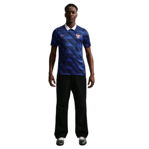 A man modeling a blue patterned France soccer jersey with a white collar, black trousers, and athletic shoes against a black background.