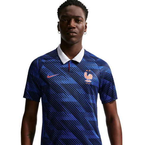 A man wearing a blue Nike France football jersey with a white collar and diagonal patterns, featuring a gold rooster crest and two stars, set against a black background.
