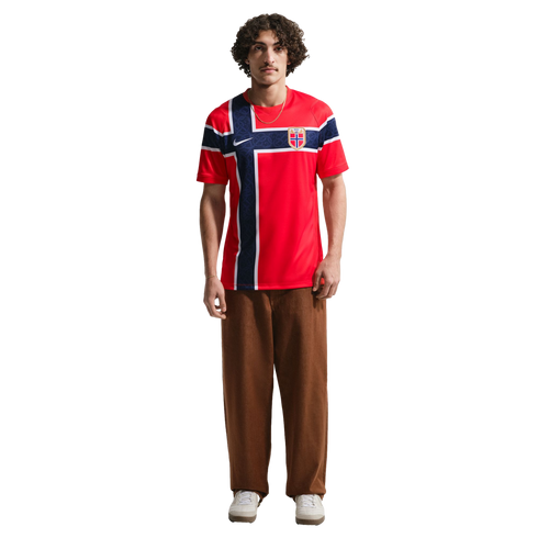 A young man stands against a black background wearing a red Nike Norway national team soccer jersey, brown baggy trousers, and white sneakers.