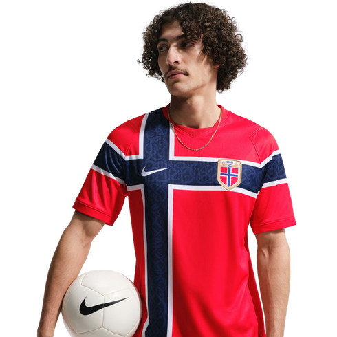 Man with curly hair wearing a red Norway national team soccer jersey and holding a white soccer ball against a black background.
