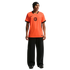 A studio photograph of a man standing against a black background. He is wearing a bright orange Netherlands national football team jersey with black trim, wide-leg black trousers, and a black beanie.