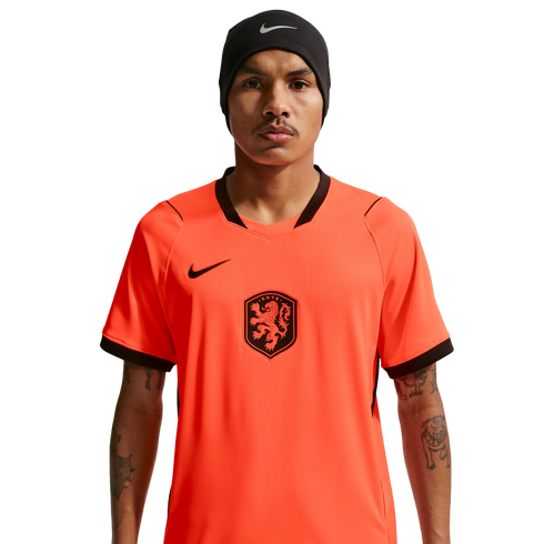A man wearing a bright orange Netherlands national football team jersey and a black Nike beanie, centered against a solid black background. He has a mustache and visible tattoos on his arms.