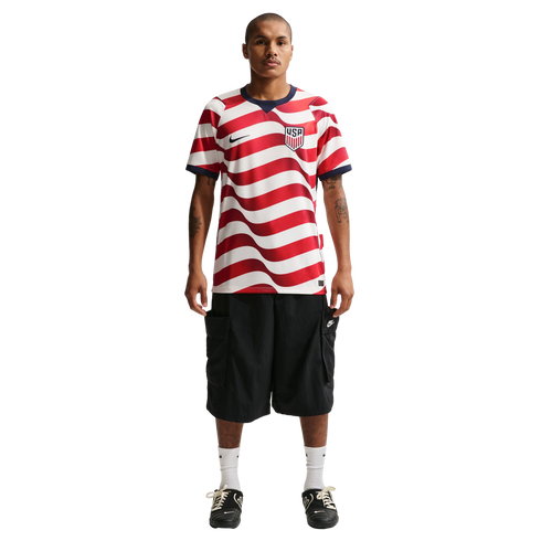Man wearing a red and white wavy-striped USA soccer jersey, black cargo shorts, and black sneakers, standing against a black background.