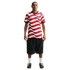 Man wearing a red and white wavy-striped USA soccer jersey, black cargo shorts, and black sneakers, standing against a black background.