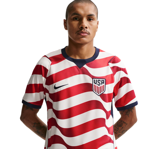 A man with a mustache in a red and white wavy-striped USA soccer jersey with a Nike logo and USA crest, against a black background. He has tattoos on his forearms.