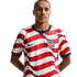 A man with a mustache in a red and white wavy-striped USA soccer jersey with a Nike logo and USA crest, against a black background. He has tattoos on his forearms.